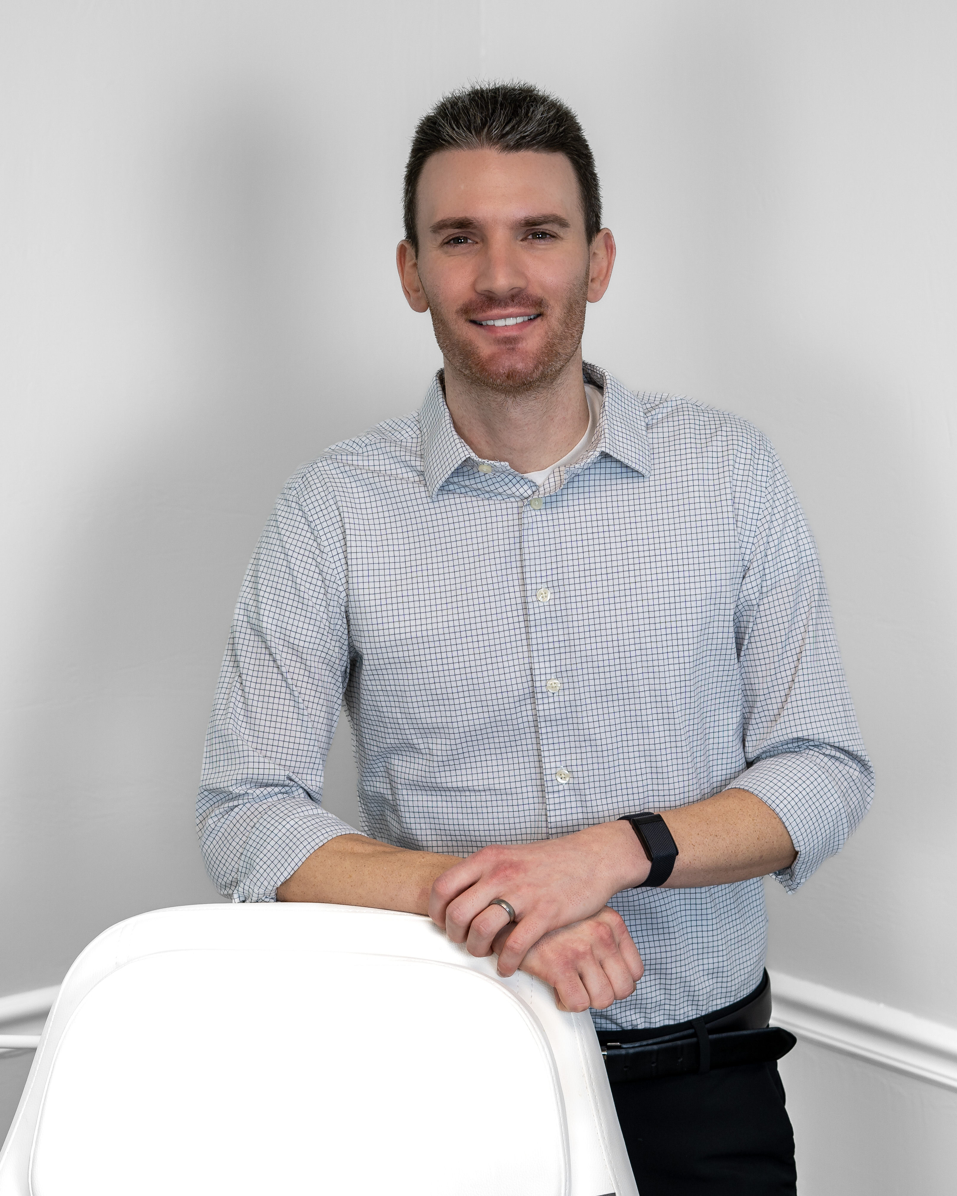 A man with dark hair and trim beard, wearing a light-colored checkered shirt, leans on a white chair, smiling at the camera.