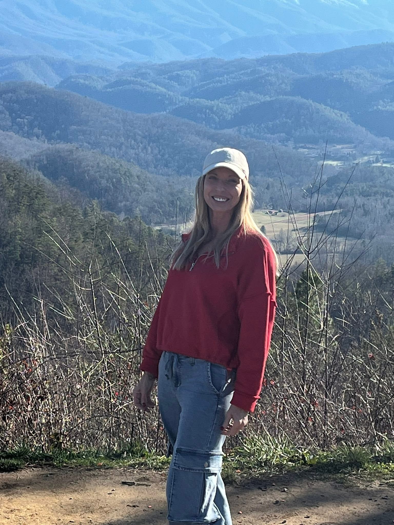 A woman with long blond hair, wearing a red sweater, jeans, and white hat, stands smiling in front of a scenic, mountainous landscape.